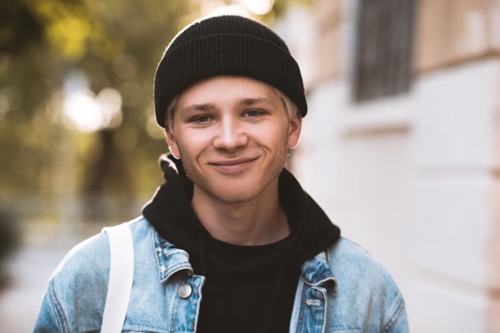 Smiling trans teen boy 16-18 year old wearing black knitted hat, hoodie and denim jacket outdoors at sun set on background. Looking at camera. 