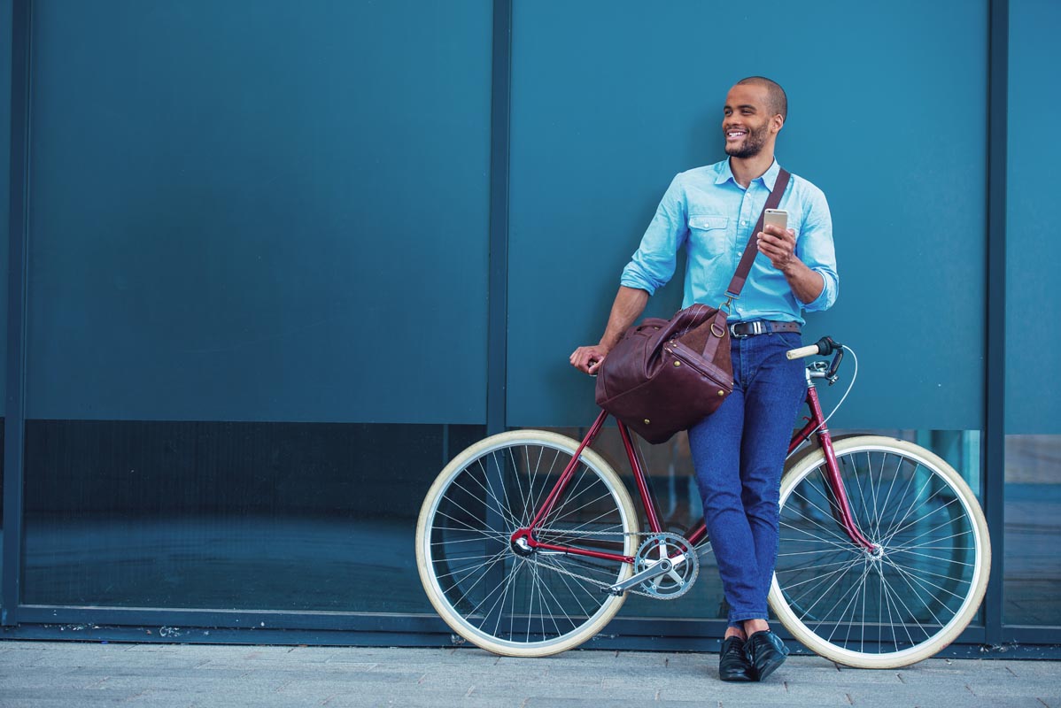 Full length portrait of handsome young African American man in casual clothes using phone, looking away and smiling while leaning on his bike, standing outdoors